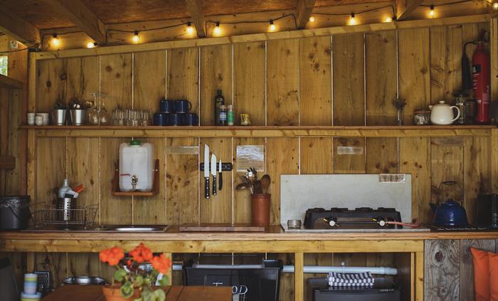A camp kitchen with festoon lighting above, a countertop with shelving above with glasses and mugs on. A fridge is below the counter and in the foregr